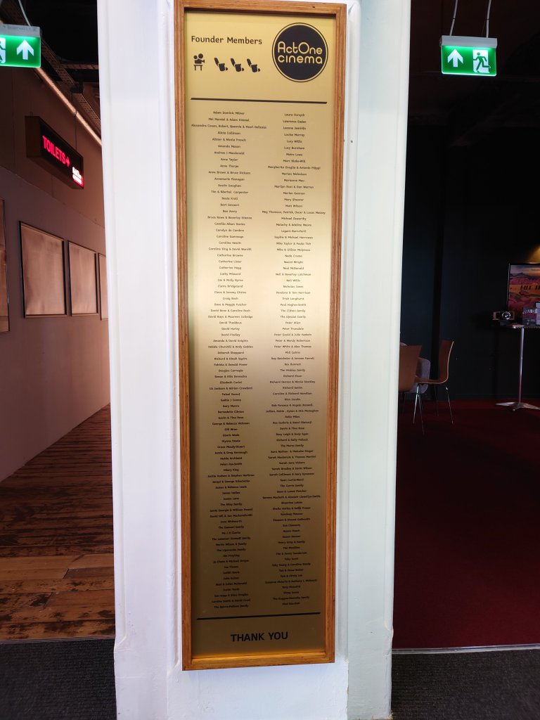 A long gold plaque with names against a white wall a sign on the left pointing to toilets and tables on the right for a seating area. The plaque is titled Founder Members Act One Cinema