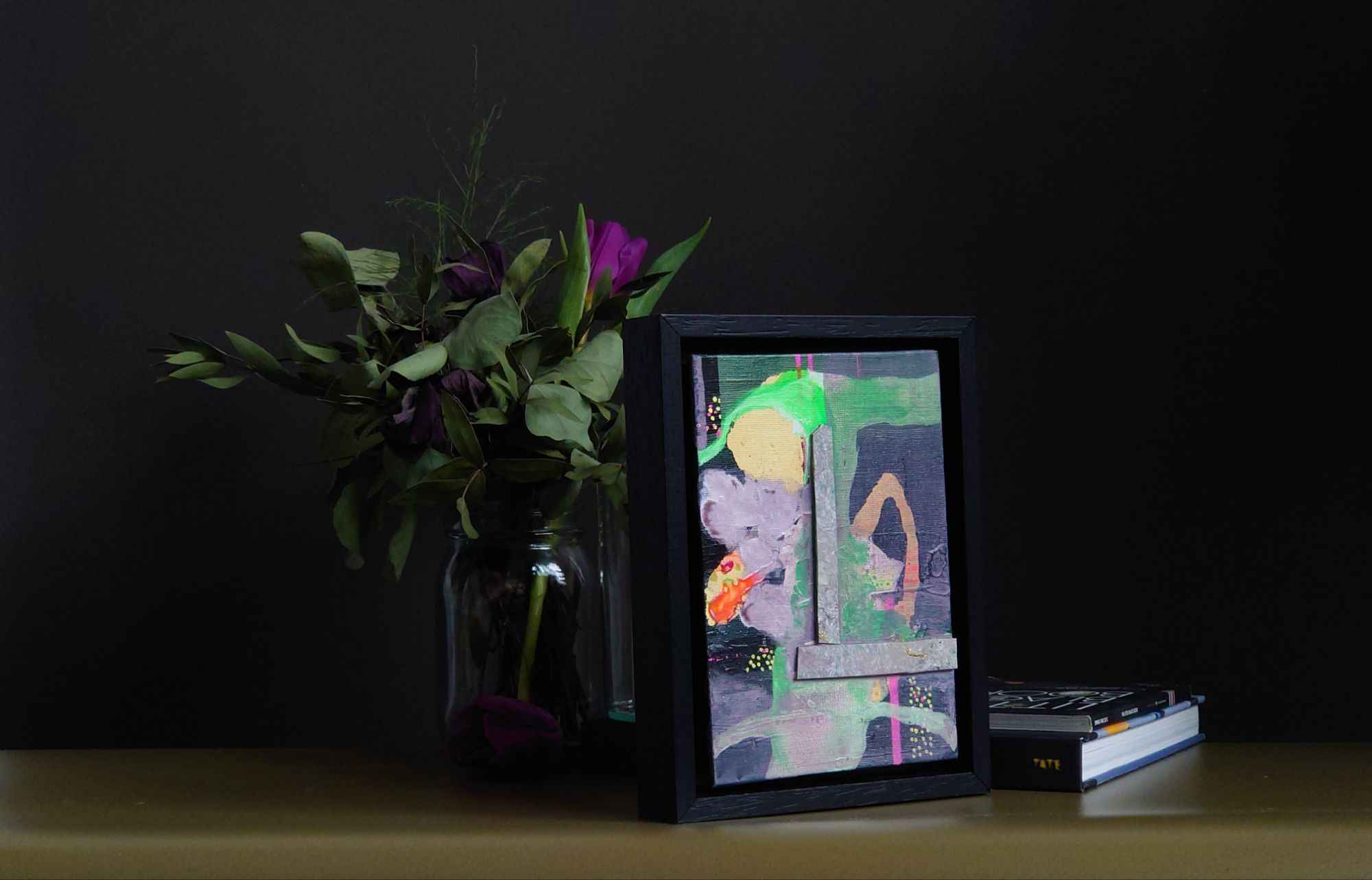 Dark grey painting with yellow red and green elements against bouquets in a vase on a shelf scene
