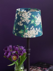 Blue and white floral lampshade on tall candlestick base against a purple background. The flowers are dainty with green sprigs. Pattern known as cherry blossom my Liberty Print