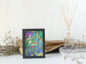 A green and purple one of a gallery of small hand painted and crafted artwork arranged with sea lavender for a whimsical scene. The frame is black and it is styled with glass reed diffuser, gold leatherette and stylish books to illustrate the artwork. 