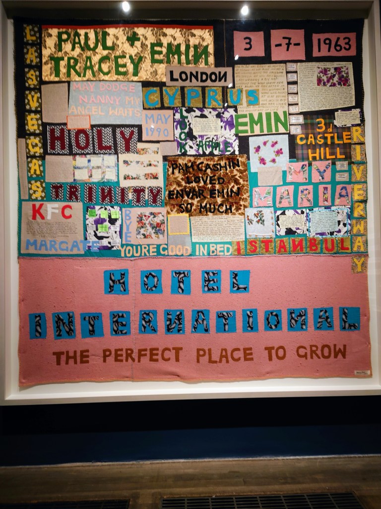 Picture of Tracy Emins artwork which is blanket with names and dates appliqued. Colours are pink, plue and gold fabric
