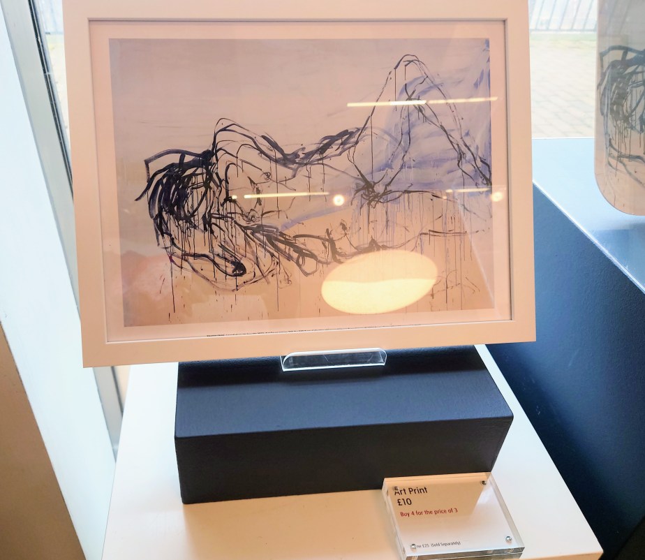 A framed abstract art print featuring flowing black lines and light colours, displayed on a white surface with a black base. A small sign indicates the price.